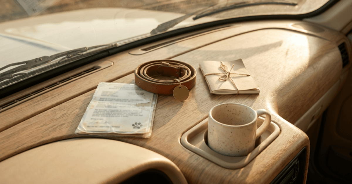 A flat-lay on a warm car dashboard of a folded laminated info card with a small paw mark, a small leather pet collar with a blank tag, a folded paper notepad, and a small ceramic mug in a cup-holder.