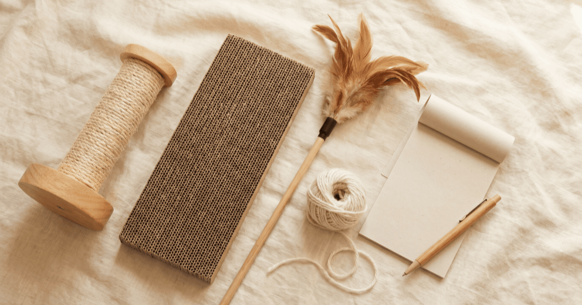 A flat-lay on warm cream linen of a cat scratcher post, a small cardboard scratch pad, a feather wand toy, a small ball of yarn, and a folded paper notepad with a pen.