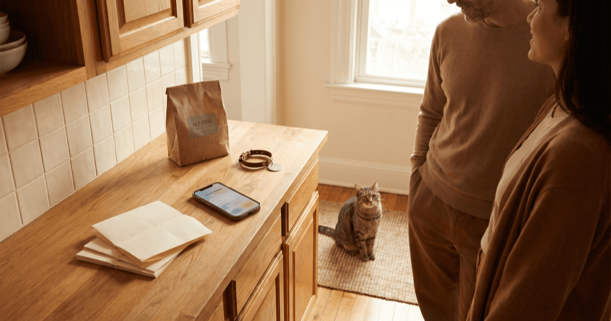 A warm sunlit kitchen counter with two adults in calm conversation, a folded paper note pad, a smartphone with a sitter-notes screen, a small pet collar with a blank tag, and a small bag of pet food on the counter.