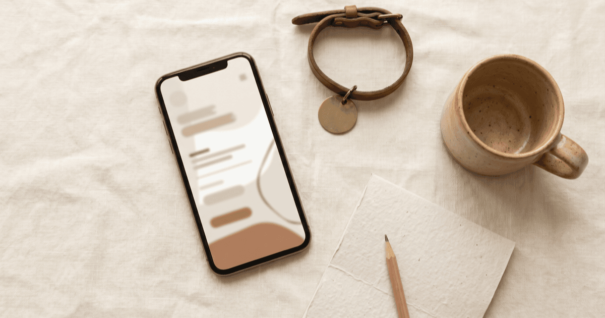 A flat-lay on warm cream linen of a smartphone showing a calm minimal account screen, a small leather pet collar with a blank tag, a folded paper notepad, and a small ceramic mug.