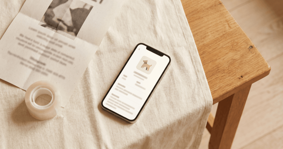 A flat-lay on warm cream linen of a smartphone with a pet profile screen, a folded printed paper lost-pet poster, and a roll of clear tape on a wooden side table.