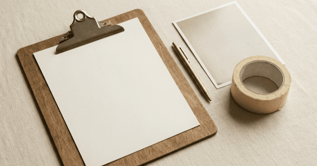 A flat-lay of a clipboard with a blank sheet of paper, a pen, a face-down printed photograph, and a roll of tape on warm cream linen.