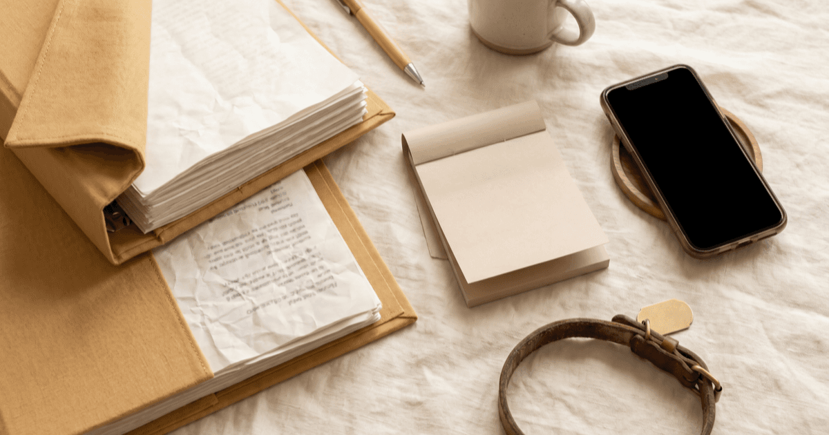 A flat-lay on warm cream linen of a thick three-ring binder slightly open with visible record sheets, a folded paper note pad, a smartphone face-down, and a small leather pet collar with a blank tag beside it.