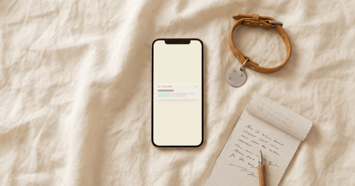 A flat-lay on warm cream linen of a smartphone showing a calm minimal alert-banner screen, a small leather pet collar with a blank QR-style tag, a folded paper notepad, and a pen.