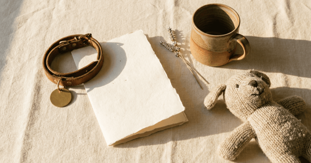 A flat-lay of a small puppy collar with a blank metal tag, a folded sheet of paper, a ceramic mug, and a soft toy on warm cream linen.