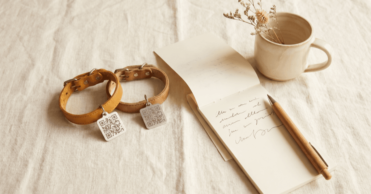A flat-lay on warm cream linen of two small leather pet collars side by side each with a blank QR-style tag, a folded paper notepad, a pen, and a small ceramic mug.