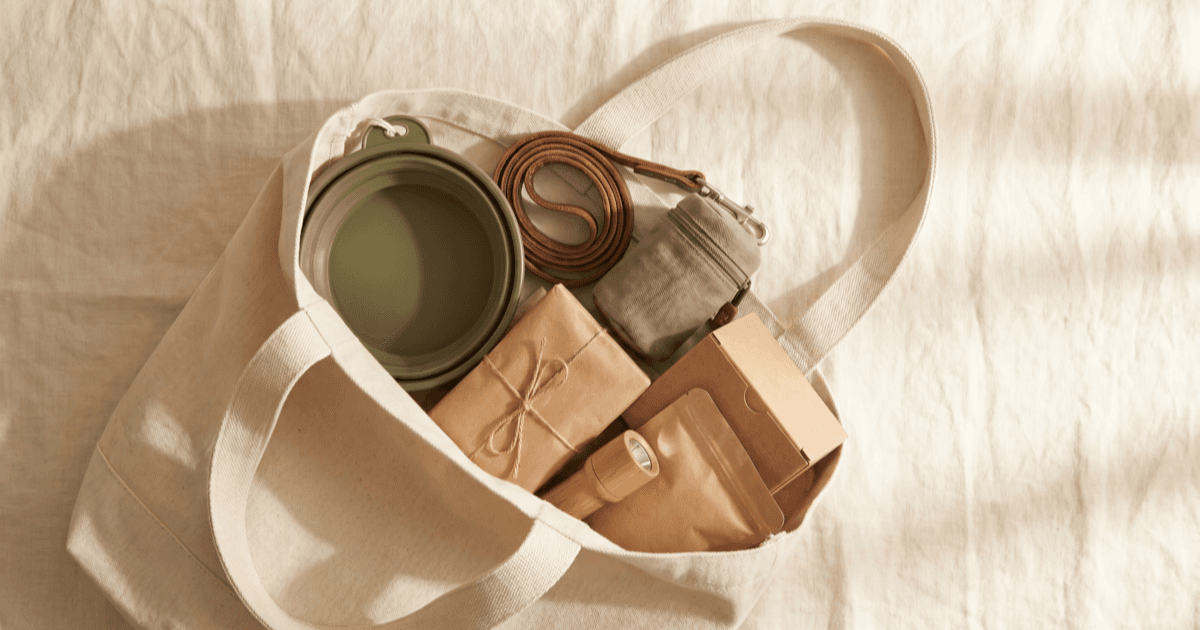 A flat-lay on warm cream linen of a canvas tote opened to show a small foldable water bowl, a coiled leash, a small carrier, a folded paper packet, a flashlight, and packaged essentials.