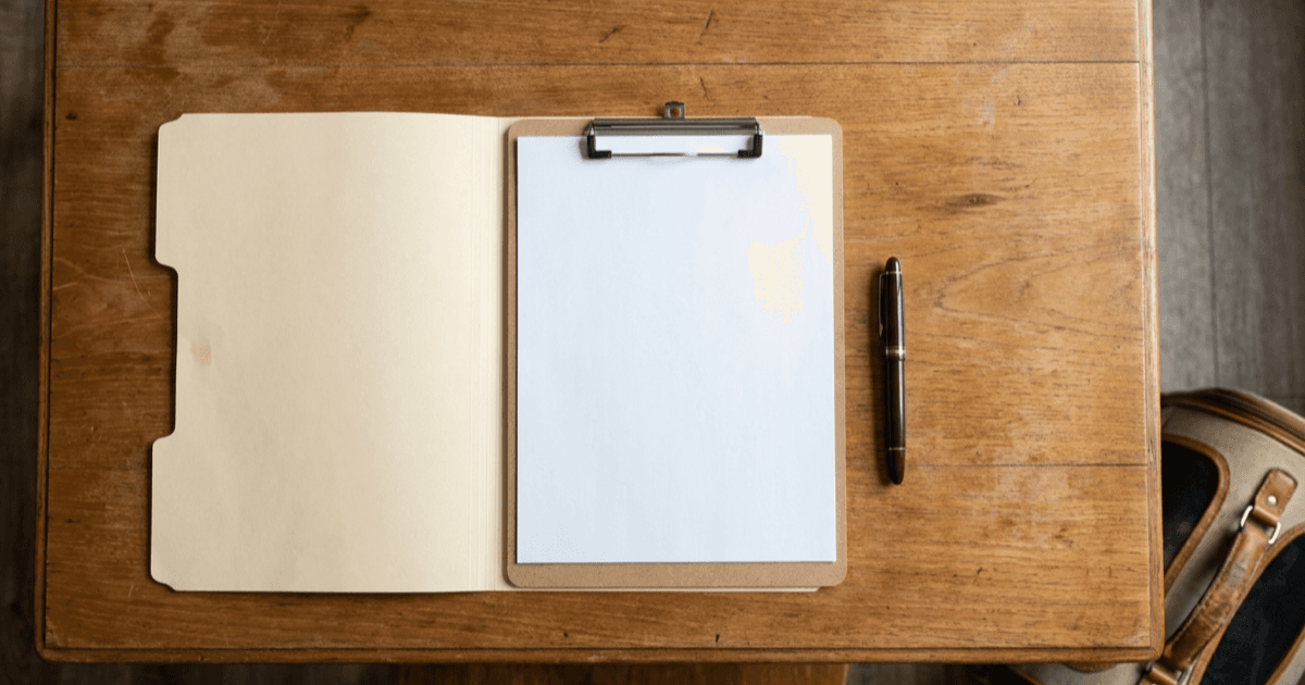 A warmly lit desk with a folded paper document, a pen, a stethoscope, and a small house plant in soft natural light.