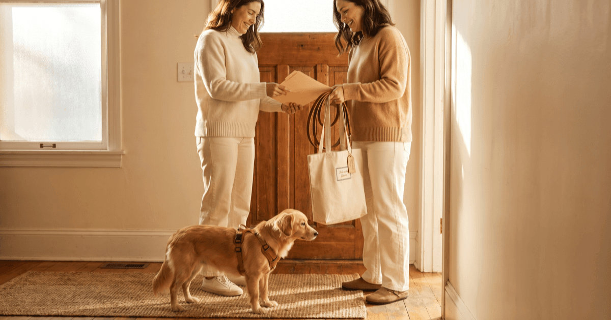A warm doorway handoff scene with two adults politely handing over a small folder of paperwork, a coiled leash, a labeled fabric tote bag, and a calm small dog standing between them in a soft sunlit entryway.
