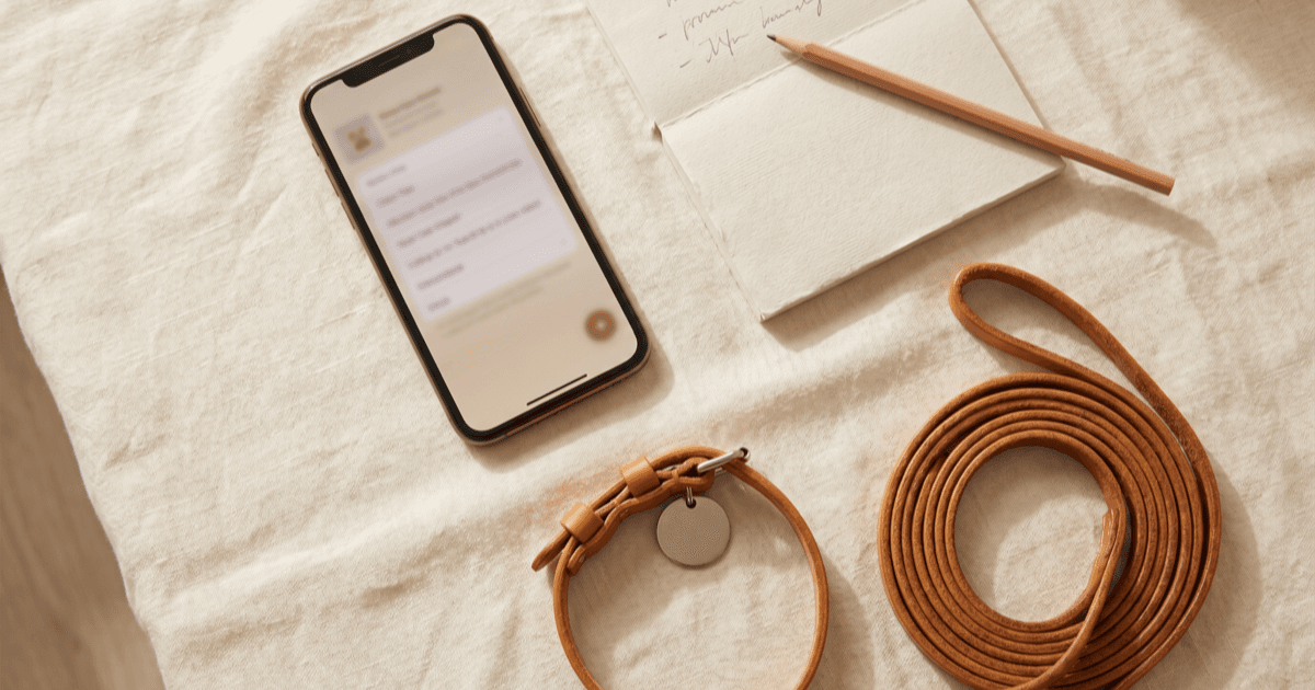 A flat-lay on warm cream linen of a smartphone with an emergency-contacts screen, a folded paper notepad with a handwritten short list, a small leather pet collar with a blank tag, and a coiled leash.