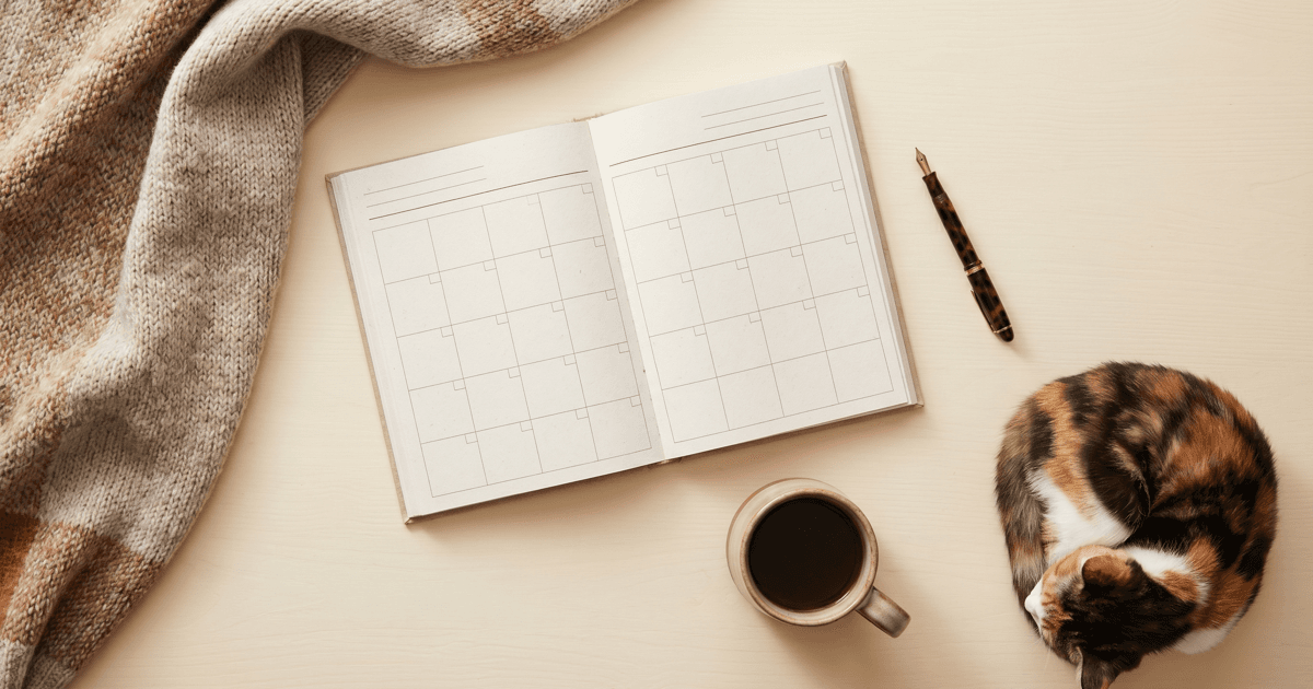 A flat-lay of a blank monthly paper planner, a pen, a mug, and a calico cat resting in the corner of the frame on a warm cream surface.
